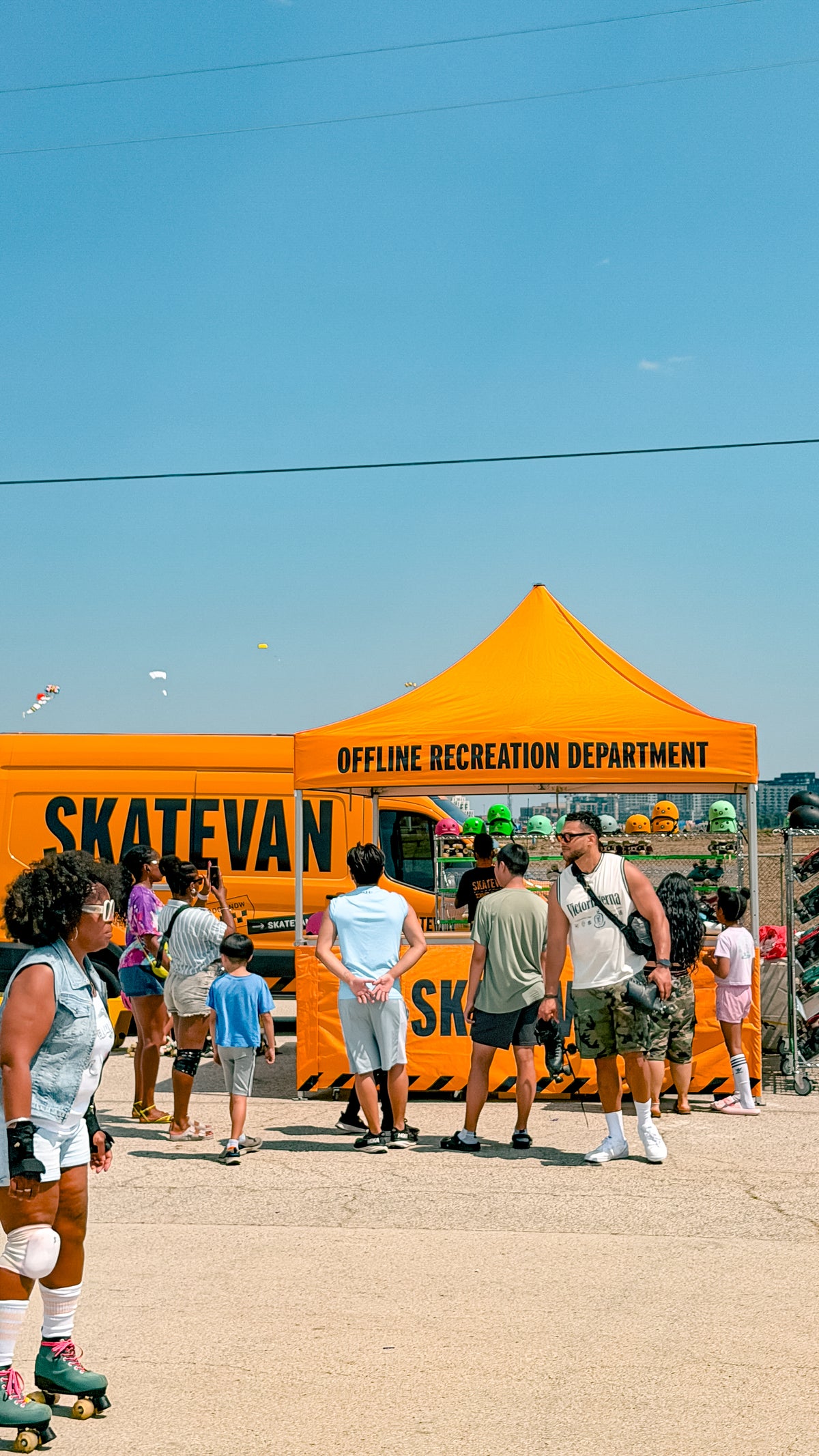 A bright orange pop-up canopy tent with 'OFFLINE RECREATION DEPARTMENT' printed in black, set outdoors on a former runway with several people standing and walking around it under a clear blue sky.